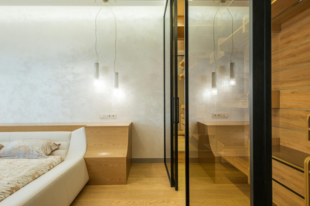 Modern bedroom with king sized bed and walk in wardrobe closet with glass doors and walls in beige and white colors lighted with led lamps