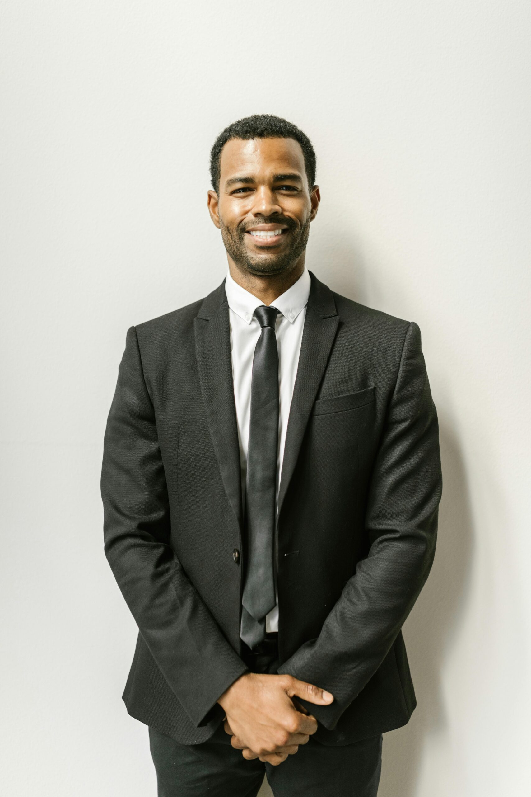 Confident man in formal attire smiling against a plain white wall.