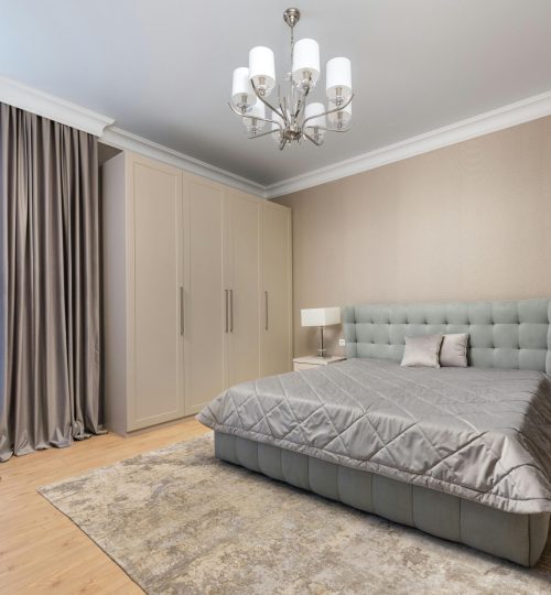 Bed with coverlet and cushions against lamps on bedside tables on parquet under chandelier in light house
